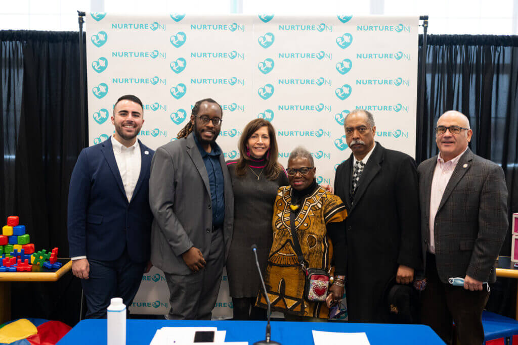 Members of the Nurture NJ Coalition — including NJ First Lady Tammy Murphy and Garden State Equality Executive Director Christian Fuscarino and Board Chair Jeannine Frisby LaRue — pose for a group photo after a press conference. They are in business formal attire, standing in front of a banner with the coalition's logo repeated as a pattern on it.